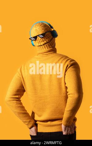 Une personne se tient debout avec le dos tourné, portant un pull tricoté jaune, un bonnet et des lunettes de soleil fraîches, avec des écouteurs bleus contre un BA jaune vif Banque D'Images