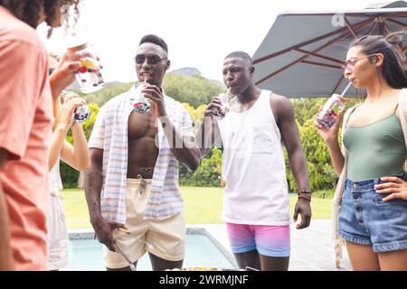 Un jeune homme afro-américain sirote un verre à côté d'une femme biraciale lors d'une fête à la piscine Banque D'Images
