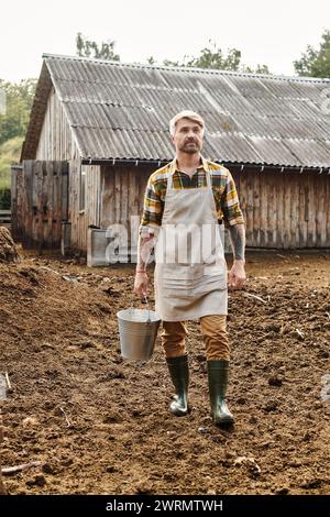 beau homme barbu avec des tatouages sur les bras tenant seau avec du lait frais alors que sur sa ferme Banque D'Images