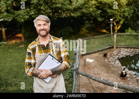 beau fermier joyeux avec des tatouages et une barbe tenant une planche à pince près de la volière et souriant à la caméra Banque D'Images