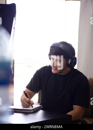 Jeune homme souriant écrit dans un cahier avec un stylo, avec des écouteurs devant son ordinateur, immergé dans le travail ou les études Banque D'Images