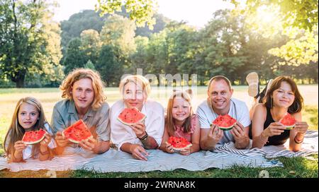 Grande famille couchée sur la couverture de pique-nique dans le parc de la ville sous l'arbre Linden pendant le week-end dimanche jour ensoleillé, souriant, riant à la caméra et mangeant l'eau Banque D'Images