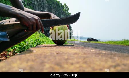 Vue d'un Africain avec une machette coupant une noix de coco, avec le tunnel de Santa Catarina comme arrière-plan à Sao Tomé, Afrique Banque D'Images