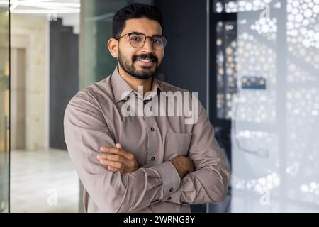 Un jeune homme professionnel avec une barbe et des lunettes, souriant en toute confiance dans un cadre de bureau contemporain bien éclairé. Banque D'Images