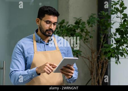 Un homme dévoué avec une barbe et des lunettes se concentre sur sa tablette tout en portant un tablier décontracté, incarnant l'entrepreneuriat et le soin des plantes. Banque D'Images