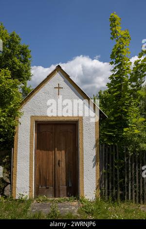 Petite maison avec une croix dans le jardin à côté de l'église de tous les Saints. Arbres frais vert vif. Ciel bleu avec nuage blanc. Dlouhomilov, Moravie, CZE Banque D'Images