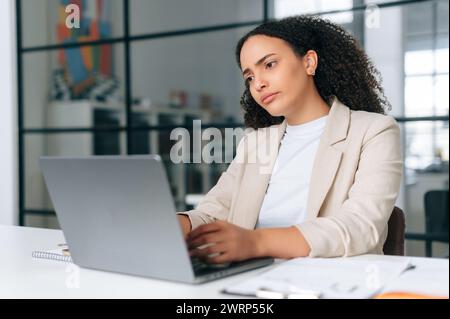 Frustré triste brésilienne ou hispanique femme d'affaires aux cheveux frisés, pdg de l'entreprise, directeur financier, est assis dans un bureau créatif moderne, contrarié à regarder un ordinateur portable, étudier des informations, lire de mauvaises nouvelles Banque D'Images