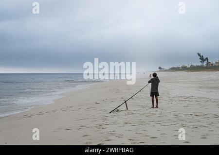 Un pêcheur préparant sa canne à poisson sur le sable blanc de la plage de Praia Seca, près des eaux bleues de l'océan Atlantique sous l'après-midi d'été ciel nuageux. Banque D'Images