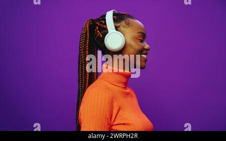 Jeune femme souriante avec des tresses bicolores violettes et des stands de maquillage à la mode dans un studio, portant des vêtements décontractés. Elle écoute de la musique avec des écouteurs, e. Banque D'Images