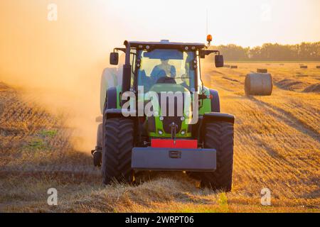 Moissonneuse-batteuse pressant la paille du champ dans les balles. Soirée d'été ensoleillée. Champ avec balles de blé pressé. Beaucoup de poussière. Tracteur avec conducteur et grosses roues conduisant sur le terrain. Agraire agricole Banque D'Images