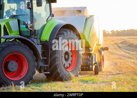 Moissonneuse-batteuse pressant la paille du champ dans les balles. Soirée d'été ensoleillée. Champ avec balles de blé pressé. Beaucoup de poussière. Tracteur avec conducteur et grosses roues conduisant sur le terrain. Agraire agricole Banque D'Images