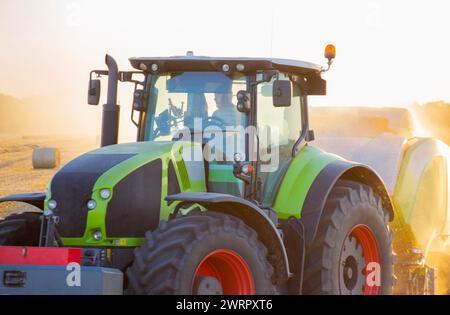 Tracteur avec conducteur et grosses roues conduisant sur le terrain. Moissonneuse-batteuse pressant la paille en balles. Soirée d'été ensoleillée. Balles de blé pressé dans les champs. Poussière. Agraire agricole. Machines agricoles Banque D'Images