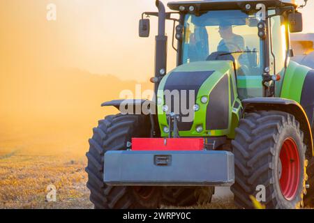 Tracteur avec conducteur et grosses roues conduisant sur le terrain. Moissonneuse-batteuse pressant la paille. Soirée d'été ensoleillée. Balles de blé pressé dans les champs. Poussière sur le terrain. Agraire agricole. Machines agricoles Banque D'Images