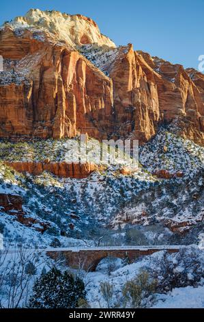 Une falaise en grès rouge doré domine un fond de canyon enneigé avec un petit pont de pierre dans le cadre inférieur au parc national de Zion, Utah. Banque D'Images