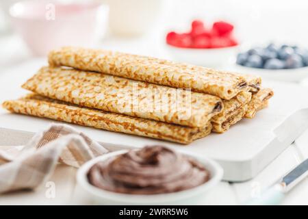 Pile de crêpes sur une assiette avec des bols de pâte à tartiner au chocolat et des baies en arrière-plan. Banque D'Images