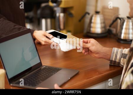 Plan recadré d'un homme payant sans contact sa commande dans un café, assis au comptoir avec un ordinateur portable, pressant la carte au terminal de point de vente, travaillant à partir de Banque D'Images