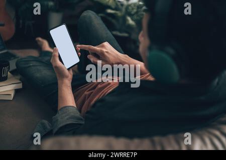 Hommes main tenant texto téléphone mobile avec écran vierge et portant casque sur canapé à la maison. Concept Relax Home. Banque D'Images