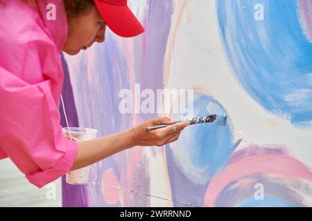 Peintre féminin dans le chapeau dessine l'image avec pinceau sur la toile pour l'exposition de rue extérieure, vue latérale rapprochée de l'artiste féminine appliquer des coups de pinceau à Banque D'Images