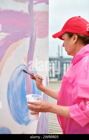 Peintre féminin dans le chapeau dessine l'image avec pinceau sur la toile pour l'exposition de rue extérieure, vue latérale rapprochée de l'artiste féminine appliquer des coups de pinceau à Banque D'Images