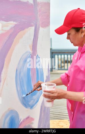 Peintre féminin dans le chapeau dessine l'image avec pinceau sur la toile pour l'exposition de rue extérieure, vue latérale rapprochée de l'artiste féminine appliquer des coups de pinceau à Banque D'Images