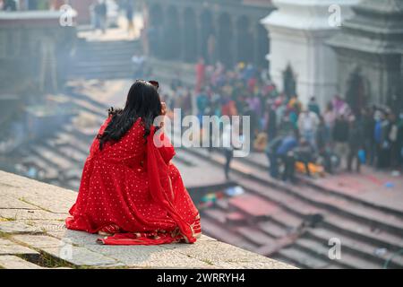 Vue arrière d'une femme en sari rouge regardant la cérémonie de crémation dans le complexe du temple de Pashupatinath, Népal, vue depuis le site de Pandra Shivalaya, symbolizi Banque D'Images