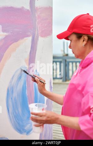 Peintre féminin dans le chapeau dessine l'image avec pinceau sur la toile pour l'exposition de rue extérieure, vue latérale rapprochée de l'artiste féminine appliquer des coups de pinceau à Banque D'Images