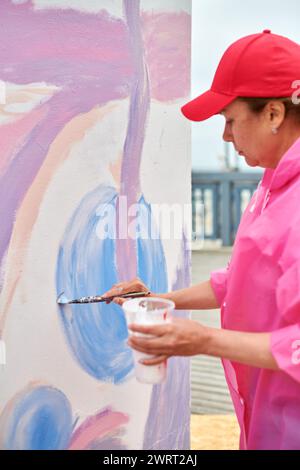 Peintre féminin dans le chapeau dessine l'image avec pinceau sur la toile pour l'exposition de rue extérieure, vue latérale rapprochée de l'artiste féminine appliquer des coups de pinceau à Banque D'Images