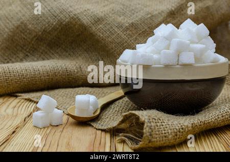 Gros plan de cubes de sucre dans un récipient en céramique sur un fond de toile de jute et une table en bois Banque D'Images