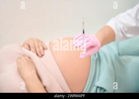 Personne en robe blanche et gants de sécurité tenant seringue avec liquide, femme enceinte couchée sur le fond, injection d'immunoglobuline RH, gynécologie et Banque D'Images