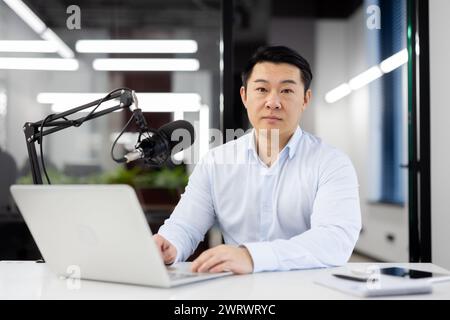 Portrait d'un jeune homme asiatique sérieux dans une chemise blanche assis dans le bureau à un bureau, travaillant avec concentration sur un ordinateur portable, regardant avec confiance l'appareil photo. Banque D'Images