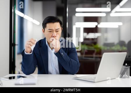 Jeune homme d'affaires asiatique assis au bureau dans le bureau et souffrant d'une crise sévère de toux et d'asthme, tenant une serviette et couvrant la bouche avec la main. Banque D'Images