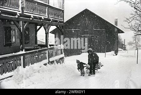 Années 1970, ...et je pensais que des vacances de ski étaient amusantes! une mère et son jeune fils tirant un petit traîneau sur la neige chargé de skis et de valise alors qu'ils tentent de se frayer un chemin à travers la neige jusqu'à leur chalet de ski. Banque D'Images