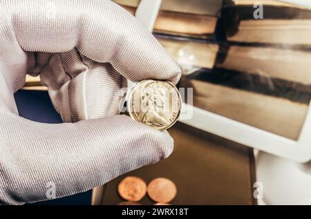 Photo de la main masculine d'une personne dans des gants blancs tenant une pièce australienne de 20 cents 1975 sur pile de livres et fond de loupe. Numismatique HO Banque D'Images