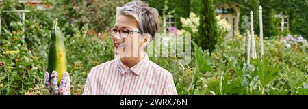 belle femme mature joyeuse dans une tenue décontractée tenant des courgettes fraîches dans son jardin, bannière Banque D'Images