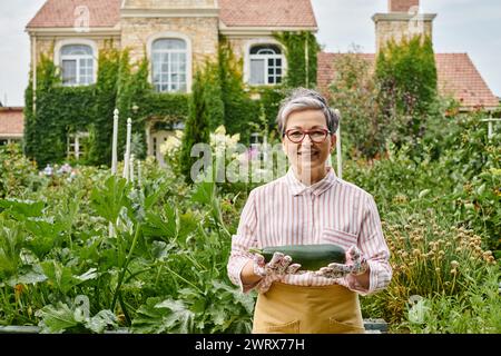 belle femme mûre joyeuse dans une tenue décontractée tenant des courgettes fraîches dans le jardin et souriant à la caméra Banque D'Images