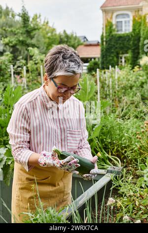 jolie femme mûre joyeuse en tenue décontractée avec des lunettes tenant des courgettes fraîches dans son jardin Banque D'Images