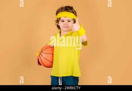 Garçon sportif en vêtements de sport avec ballon de basket-ball montrant le pouce vers le haut. Fitness, entraînement et mode de vie sain. Sport pour enfants. Petit joueur de basket-ball Banque D'Images