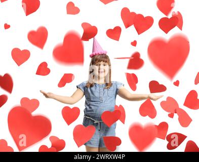 Petite fille excitée avec un chapeau d'anniversaire sous des confettis de coeur rouge isolé sur fond blanc Banque D'Images