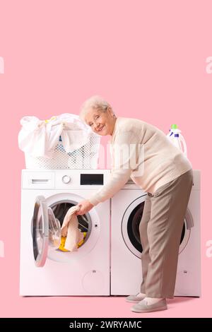 Femme âgée heureuse avec panier de vêtements mettant le linge dans la machine à laver sur fond rose Banque D'Images