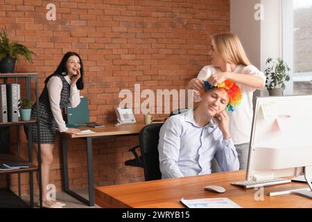 Jeune femme mettant une perruque de clown sur son collègue endormi au bureau. Célébration du jour des fous d'avril Banque D'Images