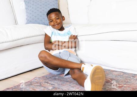 Un jeune garçon afro-américain est assis les jambes croisées sur le sol, souriant, à la maison Banque D'Images