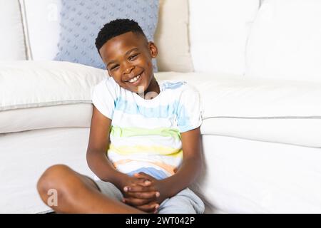 Un jeune garçon afro-américain est assis les jambes croisées sur un canapé, souriant, à la maison Banque D'Images