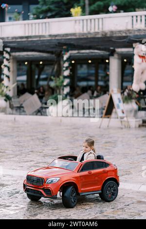 Petite fille souriante conduit une voiture rouge jouet sur la place Banque D'Images