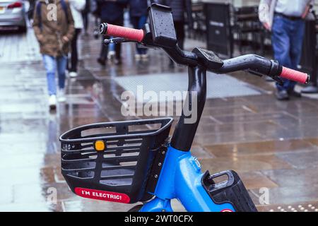 E-bike de location Blue Dott avec panier noir et signe I’m Electric garé sur une rue humide. Les gens marchant en arrière-plan. Centre de Londres, Angleterre. Banque D'Images