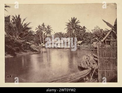 Village situé sur un virage dans une rivière. Inconnu, photographe des années 1850–1890 vue d'un village situé dans un virage d'une rivière. Les bâtiments au toit de chaume, certains sur pilotis et d'autres avec des clôtures en bois, se trouvent au bord de l'eau, entourés de grands palmiers et d'autres végétaux tropicaux. Des bateaux de différentes tailles et formes sont attachés le long de la rive. Plusieurs bateaux au premier plan sont chargés de poteaux en bois. Banque D'Images