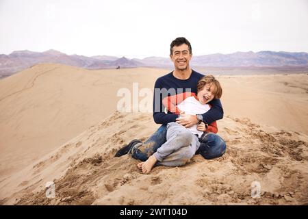 Portrait d'homme heureux jouant avec son fils assis sur une dune de sable Banque D'Images