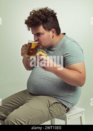 Homme, boisson et bière avec hamburger en studio pour une alimentation malsaine, restauration rapide et nutrition sur chaise. Plus de taille, affamé et homme personne avec de l'alcool sur Banque D'Images