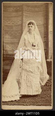 Portrait d'une mariée. Inconnu, photographe vers 1880–1890 Portrait d'une mariée portant un voile sur toute la longueur du sol. Elle tient un ventilateur dans une main à sa taille. La jupe de sa robe a des niveaux avec un motif floral ou feuille et l'ourlet est plissé. Banque D'Images