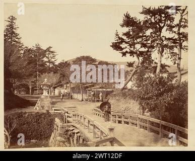 Homme debout sur un pont. Inconnu, photographe des années 1850–1890 vue d’un homme debout à une extrémité d’un pont menant à un petit village. L'homme s'appuie contre un poteau de bois dans la rambarde du pont et tient un parasol, ou parapluie en papier d'huile, sur une épaule. Un tachidoro, ou lanterne sur piédestal en pierre, se trouve à l'extrémité gauche du pont et comporte des inscriptions japonaises sur une partie de la base. Plusieurs personnes peuvent être vues se déplaçant sur le chemin de terre qui court du pont à travers le village. Banque D'Images