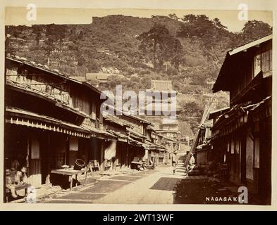 Nagasaki. Attribué à Felice Beato, photographe (anglais, né en Italie, 1832 - 1909) Baron Raimund von Stillfried, imprimeur (autrichien, 1839 - 1911) 1860 vue en bas d'une rue menant au temple Kofuku-ji à Kojiyamachi, Nagasaki. La rue est bordée des deux côtés de petites boutiques, et mène à une petite montagne parsemée de maisons. Deux personnages se tiennent dans la rue face à face dans la conversation, et deux enfants sont assis sur une petite plate-forme devant un magasin au premier plan sur le côté gauche de l'image. (Recto, impression) en bas à droite, écrit à la main en négatif : 'NAGASAKI'. Banque D'Images